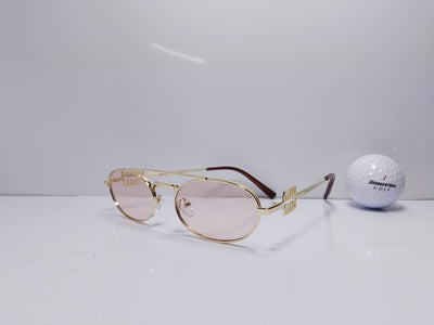 Gold-framed sunglasses with pink lenses on a white surface next to a golf ball.